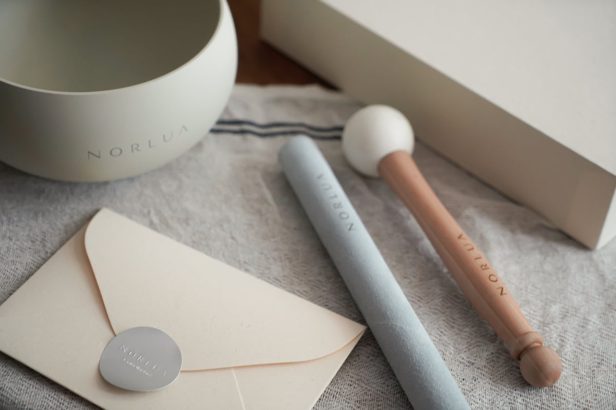 NORLUA brass singing bowl with Playing stick, Wooden mallet, envelope and Protective gift box on textured fabric.