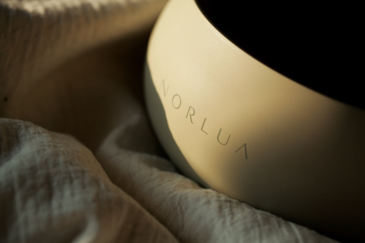Close-up of the NORLUA brass singing bowl logo and matte surface beside softly folded fabric.