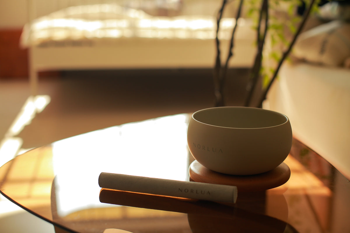 NORLUA brass singing bowl with Resting base and Playing stick on a glass table in warm sunlight.